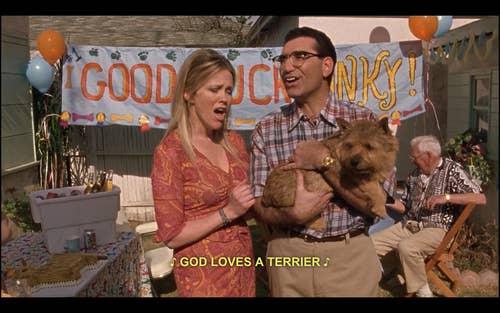 A still from the movie Best in Show, where Catherine O'Hara and Eugene Levy are holding a dog at some kind of yard party. They're singing, and the caption reads: "God loves a terrier"