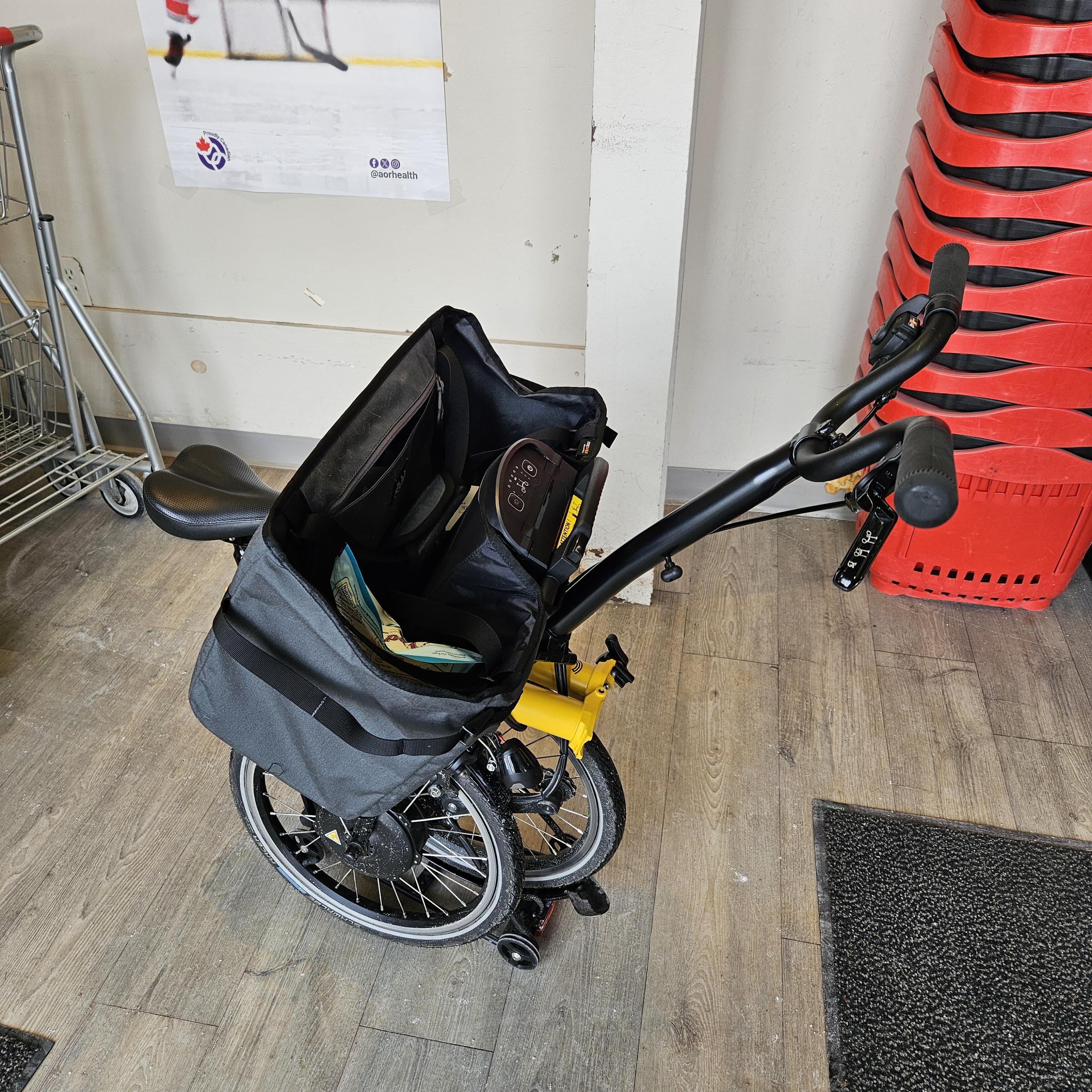 A yellow electric brompton folded into a "shopping cart". The back wheel is folded under so that it rests on the rear rack, where there are little wheels just for this purpose. The front wheel is folded to the side and back so that it's next to the rear wheel, off the ground. The seat is down so that the seat post holds the wheels in place, it won't unfold accidentally. The handlebars are up to act as a shopping cart handle. There is a basket on the luggage block, which is now swung around to be between the seat and the handlebars. The bike is next to shopping carts and baskets in a grocery store. I have a few items in the basket.