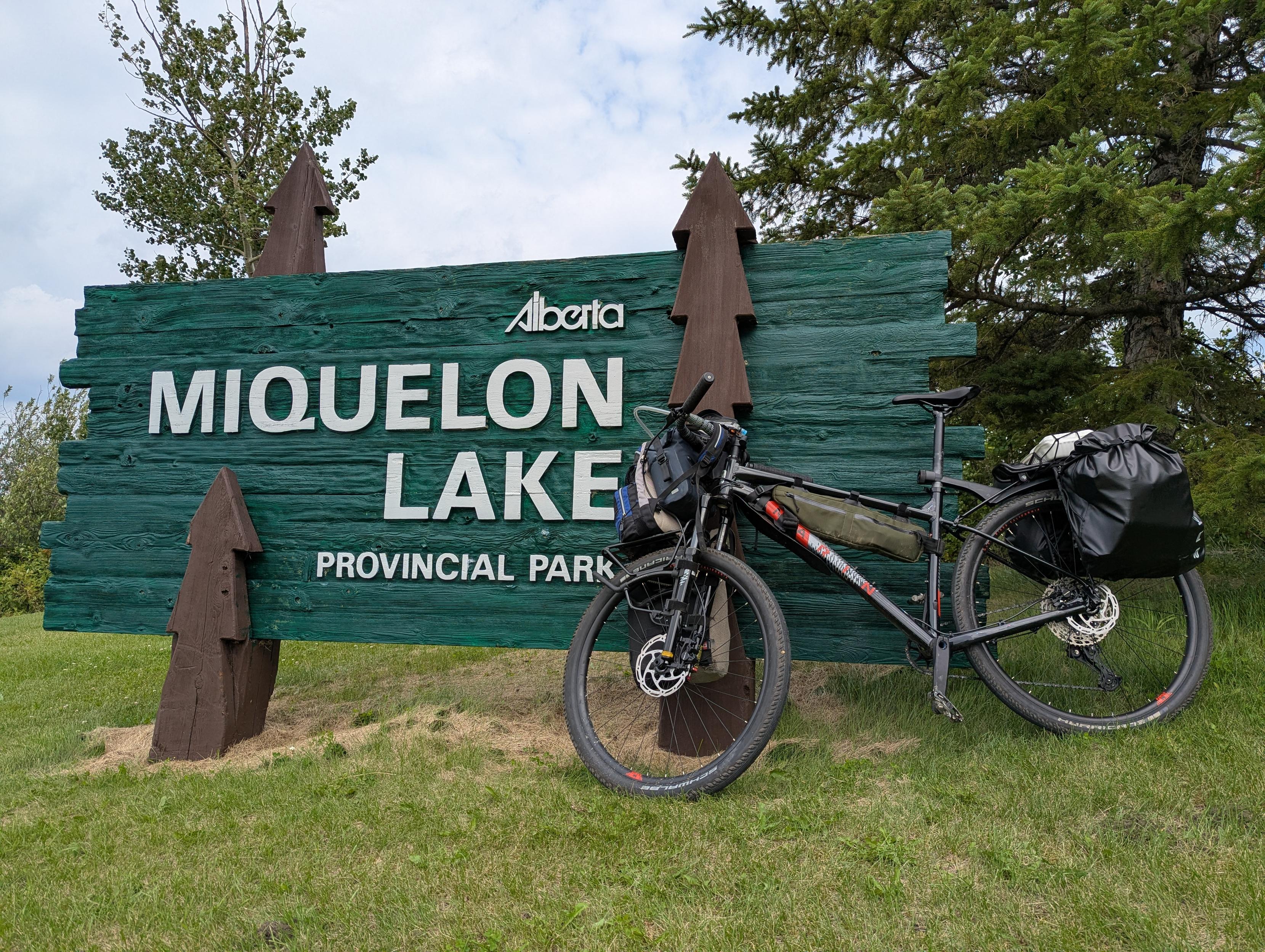 A photo of a laden mountain bike leaning against a wooden green sign that reads:
Alberta
Miquelon Lake
Provincial Park