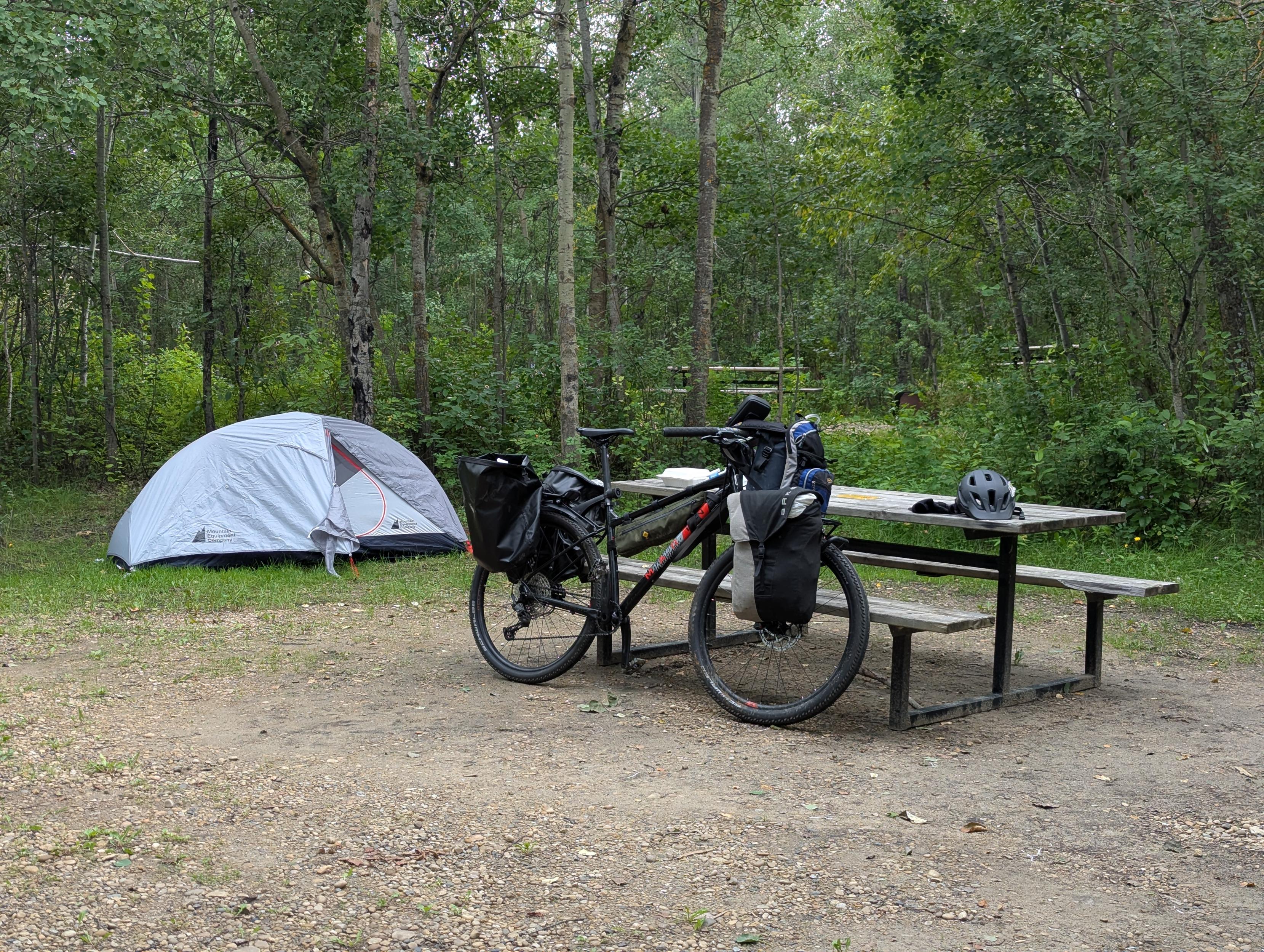 A photo of a laden mountain bike leaning against a picnic table at a campsite in a park. There is a small one-person tent set up near some trees.